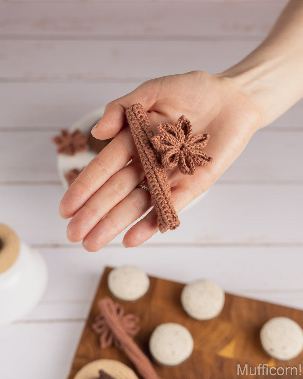A hand holds a small crocheted brown cinnamon stick and star anise hair clip against a light wood background, with more matching clips and decorative props blurred in the foreground and background.
