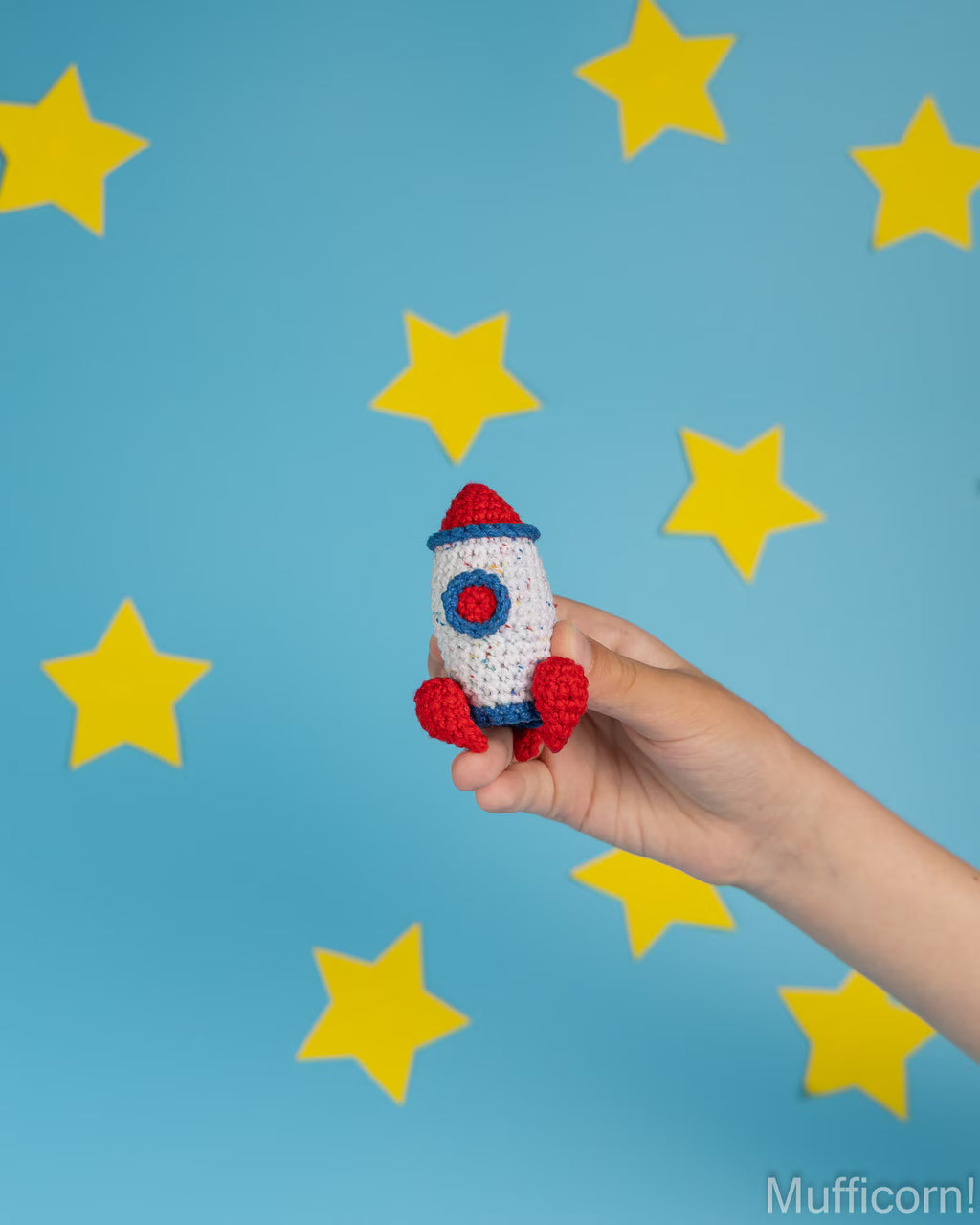A hand holds a small crocheted rocket toy against a bright blue background decorated with yellow star cutouts. The rocket is white with red and blue accents.