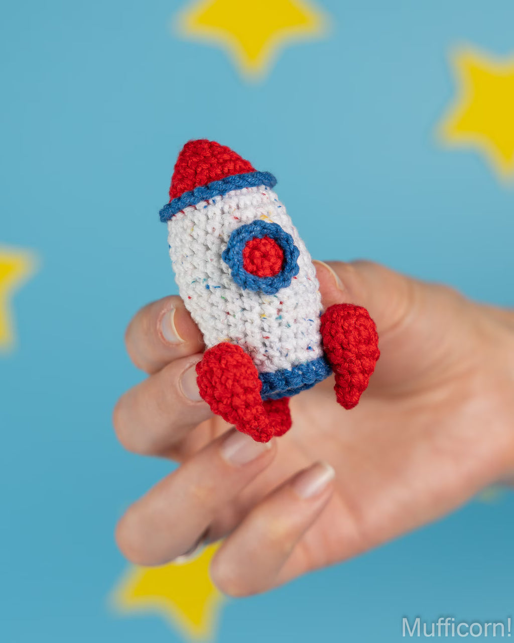 A hand holding a small crocheted rocket toy in red, white, and blue, set against a bright blue background with yellow star shapes.