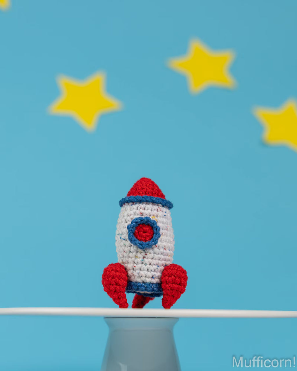 A crocheted toy rocket in red, white, and blue sits on a white surface against a bright blue background with yellow star shapes.