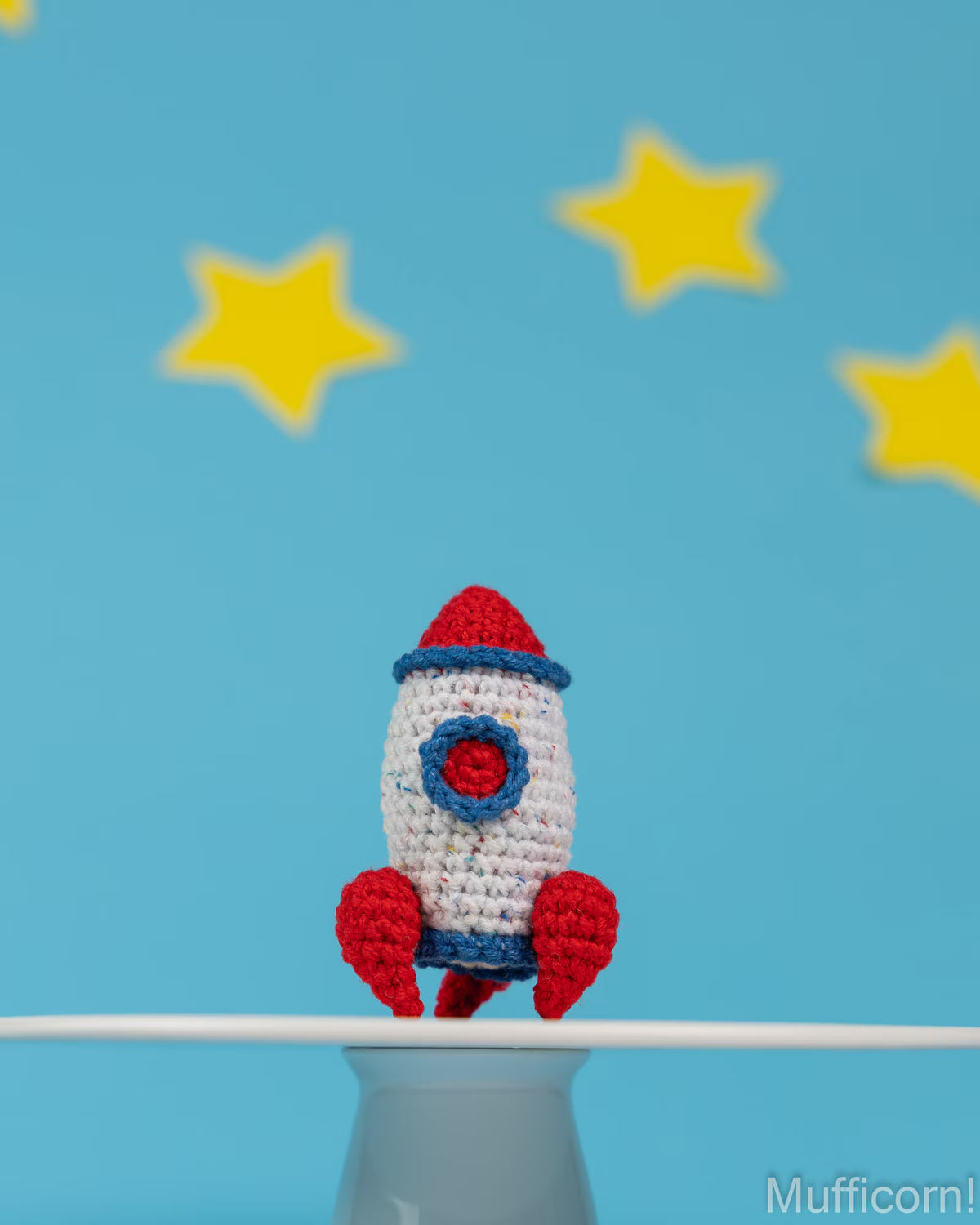 A crocheted toy rocket in red, white, and blue sits on a white surface against a bright blue background with yellow star shapes.