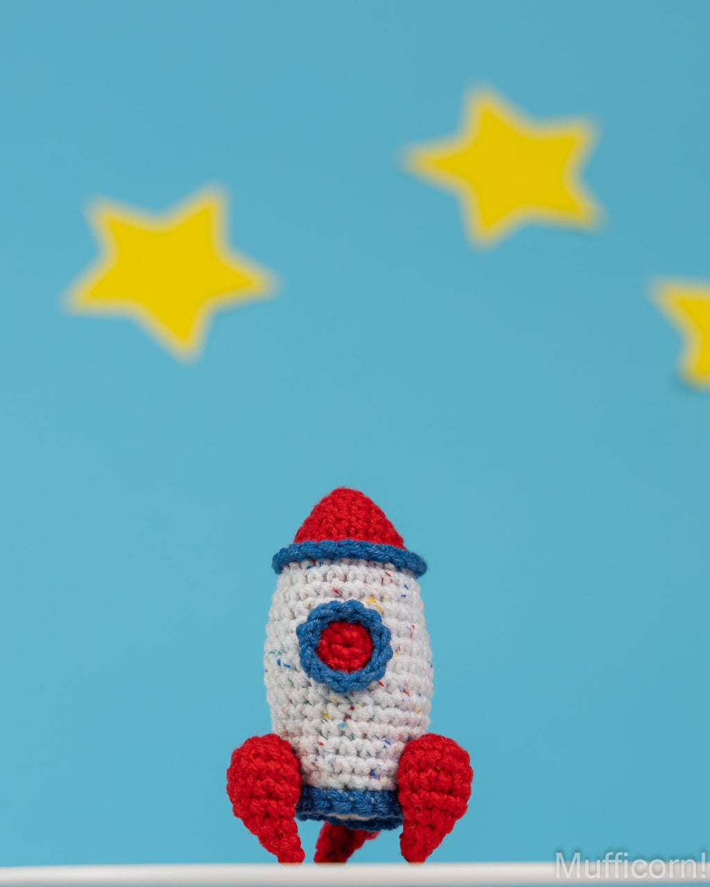 A crocheted toy rocket in red, white, and blue sits on a white surface against a bright blue background with yellow stars.