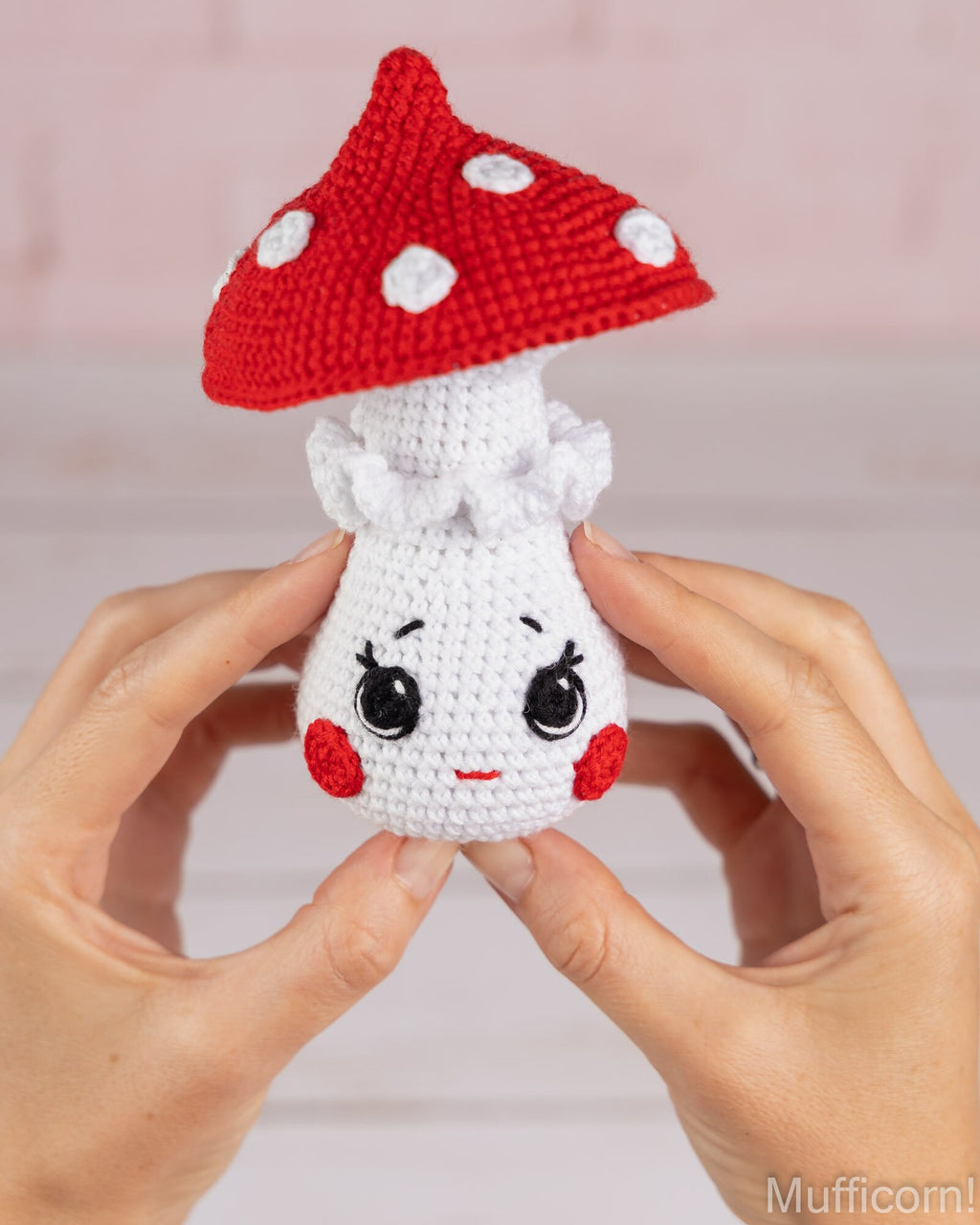 A small crocheted mushroom character being held between two hands. The mushroom has a red cap with white polka dots, a white stem and face, and black embroidered eyes and mouth.