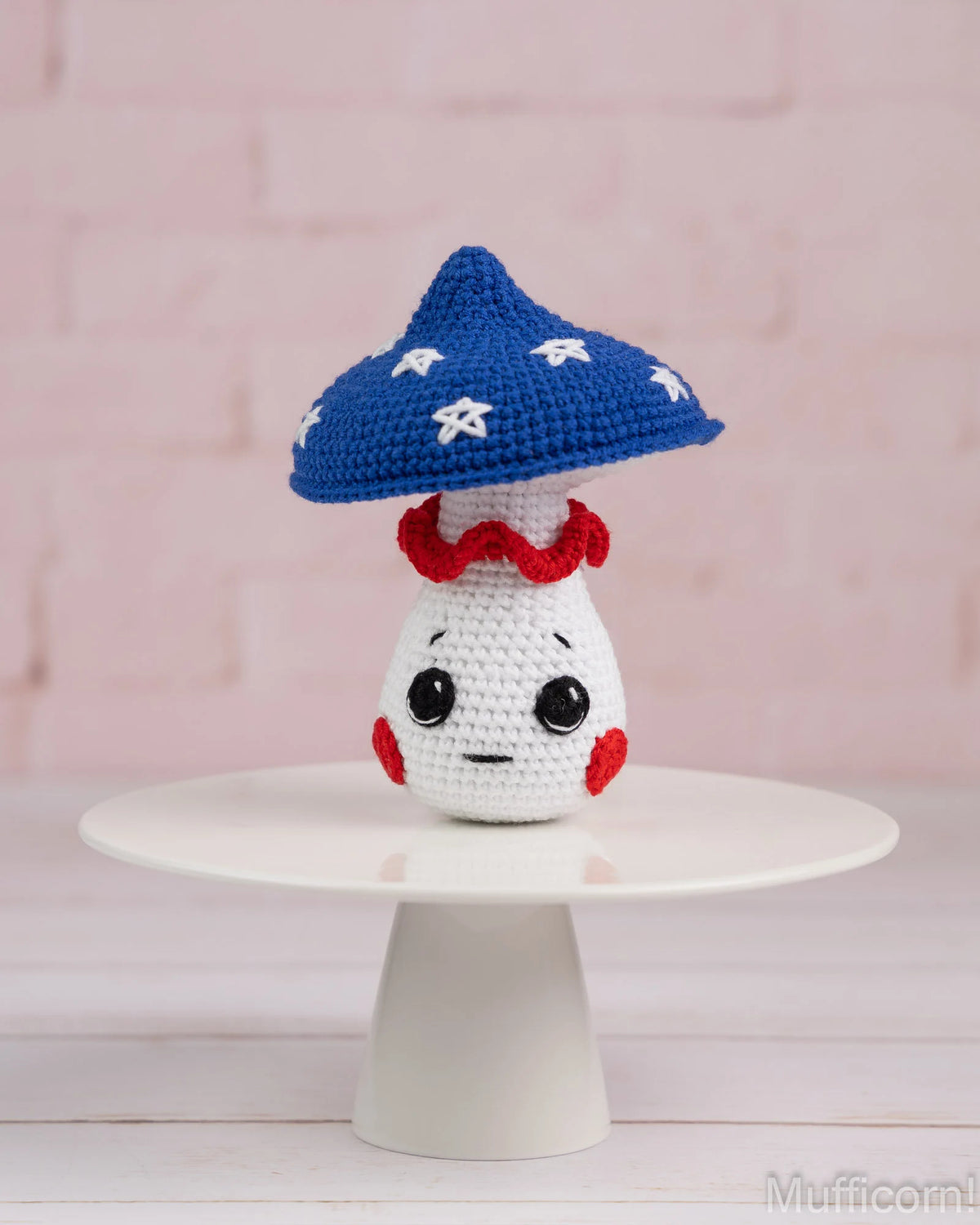 A crocheted mushroom character toy with a blue cap, white body, red ruffled collar, and black facial features, displayed on a white pedestal against a pink brick wall background.