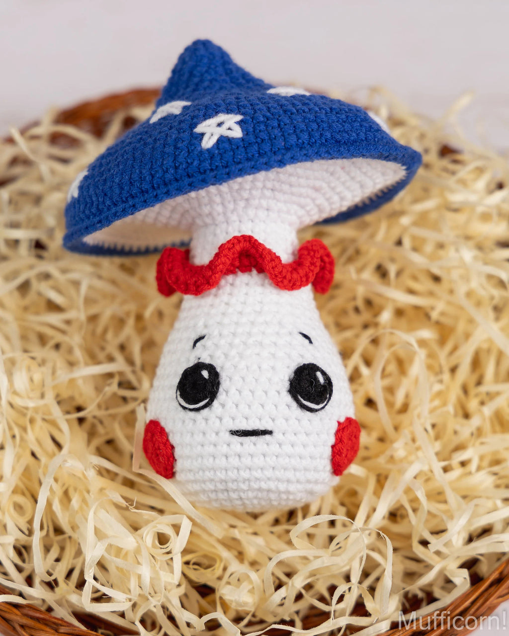 A crocheted mushroom character toy with a blue cap, white body, red ruffled collar, and embroidered face, displayed on a bed of beige shredded paper.