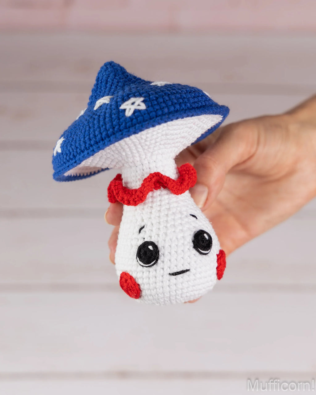 A hand holding a crocheted mushroom character toy with a blue cap, white body, red ruffled collar, and embroidered face against a soft light background.