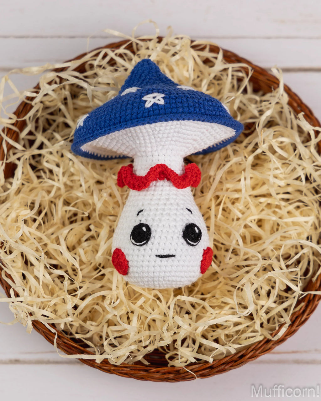 A crocheted mushroom character toy with a blue cap, white body, red collar and cheeks, and black embroidered eyes and mouth, displayed in a wicker tray filled with shredded straw on a white wood background.