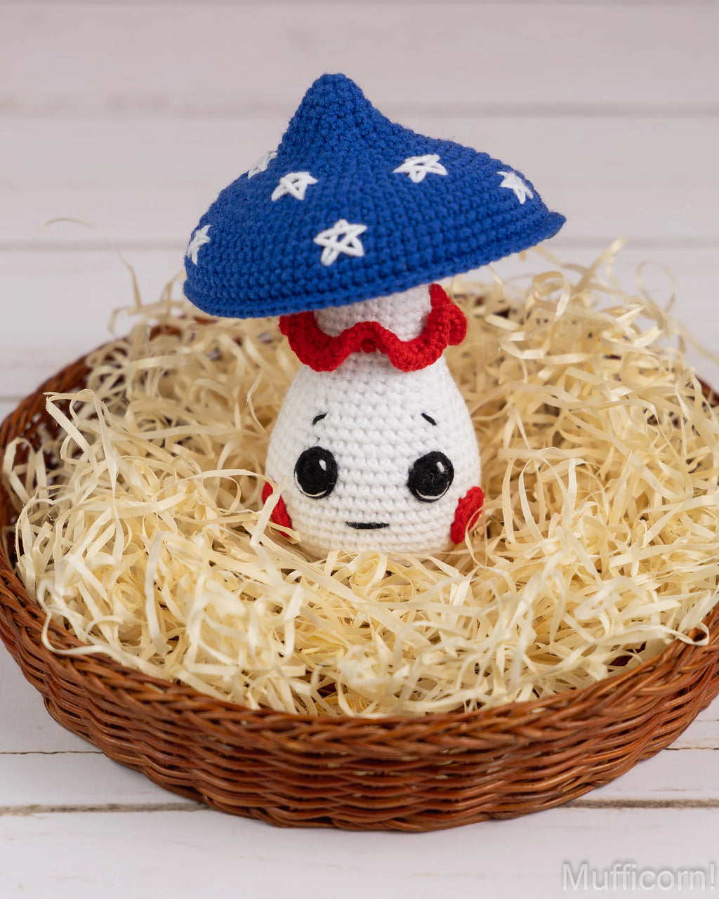 A crocheted mushroom character toy with a blue cap, white body, and red ruffled collar, sitting in a wicker basket filled with shredded paper on a white wood background.