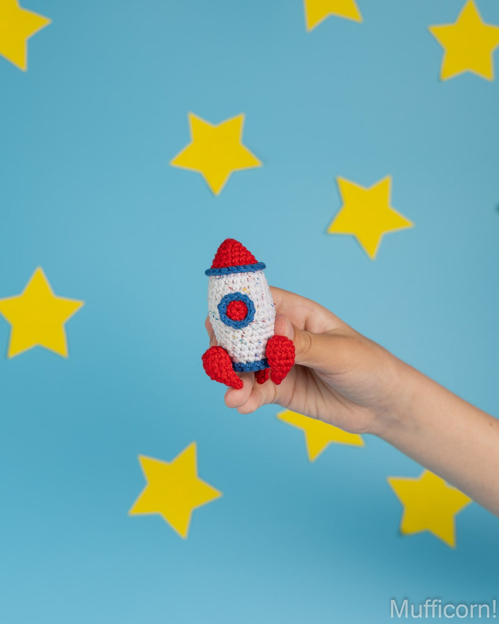 A hand holds a small crocheted rocket toy against a bright blue background decorated with yellow star cutouts. The rocket is white with red and blue details.