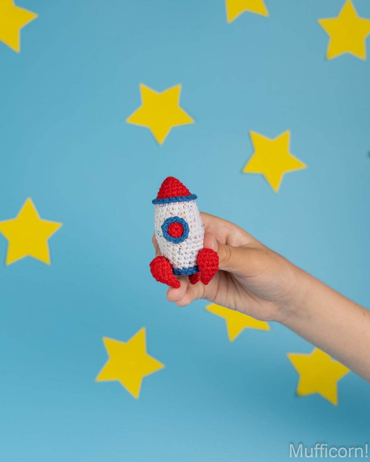 A hand holds a small crocheted rocket toy against a bright blue background decorated with yellow star cutouts. The rocket is white with red and blue details.