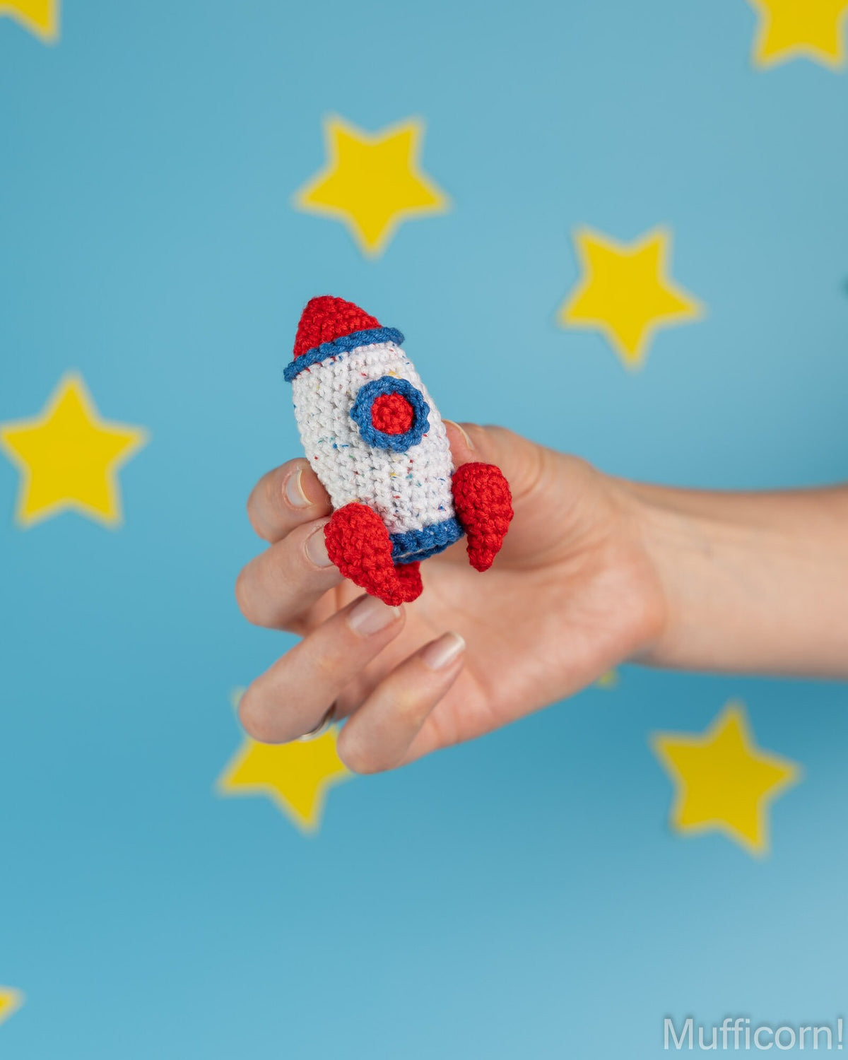 A hand holding a small crocheted rocket toy in red, white, and blue, set against a light blue background with yellow star shapes.