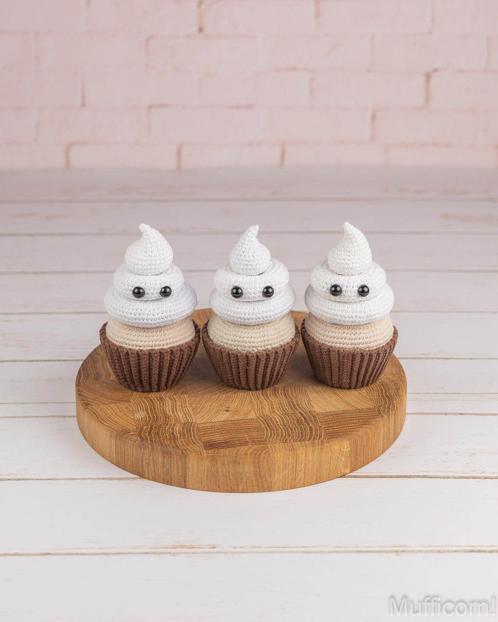 Three small crocheted ghost cupcakes with white and beige frosting-like tops, brown wrappers, and black eyes, displayed on a round wooden board against a pale pink brick wall and white wood surface.
