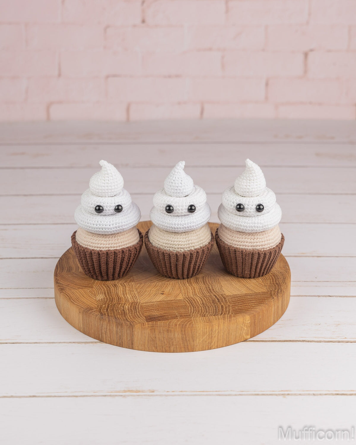Three small crocheted ghost cupcakes with white and beige frosting-like tops, brown wrappers, and black eyes, displayed on a round wooden board against a pale pink brick wall and white wood surface.