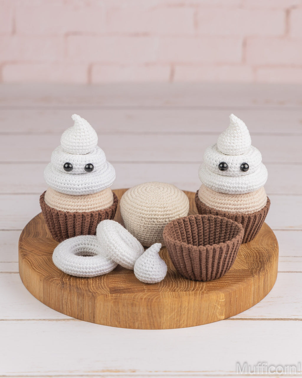 A set of crocheted cupcake toys and accessories displayed on a round wooden tray against a light pink brick wall and white wood floor. The set includes two white and beige crocheted cupcakes with brown wrappers, a matching beige cupcake topper, a brown cupcake wrapper, a white ring topper, and a small white kiss-shaped piece.