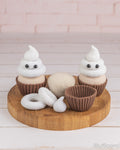 A set of crocheted cupcake toys and accessories displayed on a round wooden tray against a light pink brick wall and white wood floor. The set includes two white and beige crocheted cupcakes with brown wrappers, a matching beige cupcake topper, a brown cupcake wrapper, a white ring topper, and a small white kiss-shaped piece.