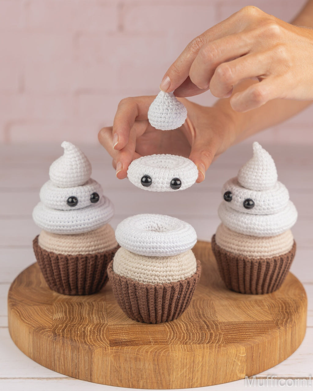 A person's hands are shown assembling a crocheted ghost-shaped cupcake, placing a small white crochet piece onto a larger white piece. Three finished cupcake toys are displayed on a round wooden board against a light pink brick wall background.