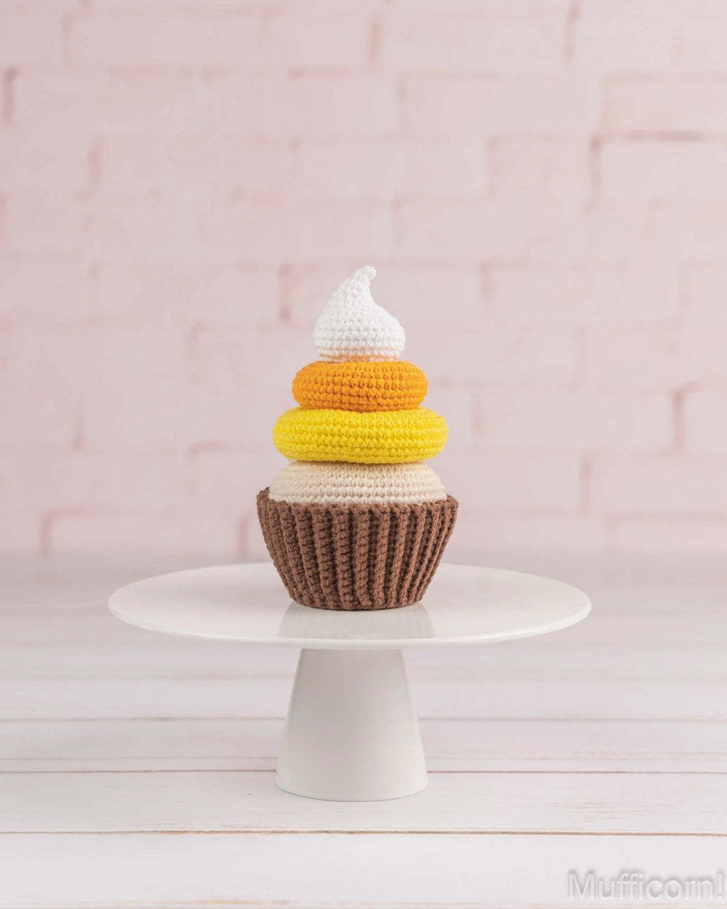 A crocheted cupcake decoration or plush toy with a brown base, yellow, orange, and cream layers, and a white frosting top, displayed on a white pedestal stand against a pale pink brick wall.
