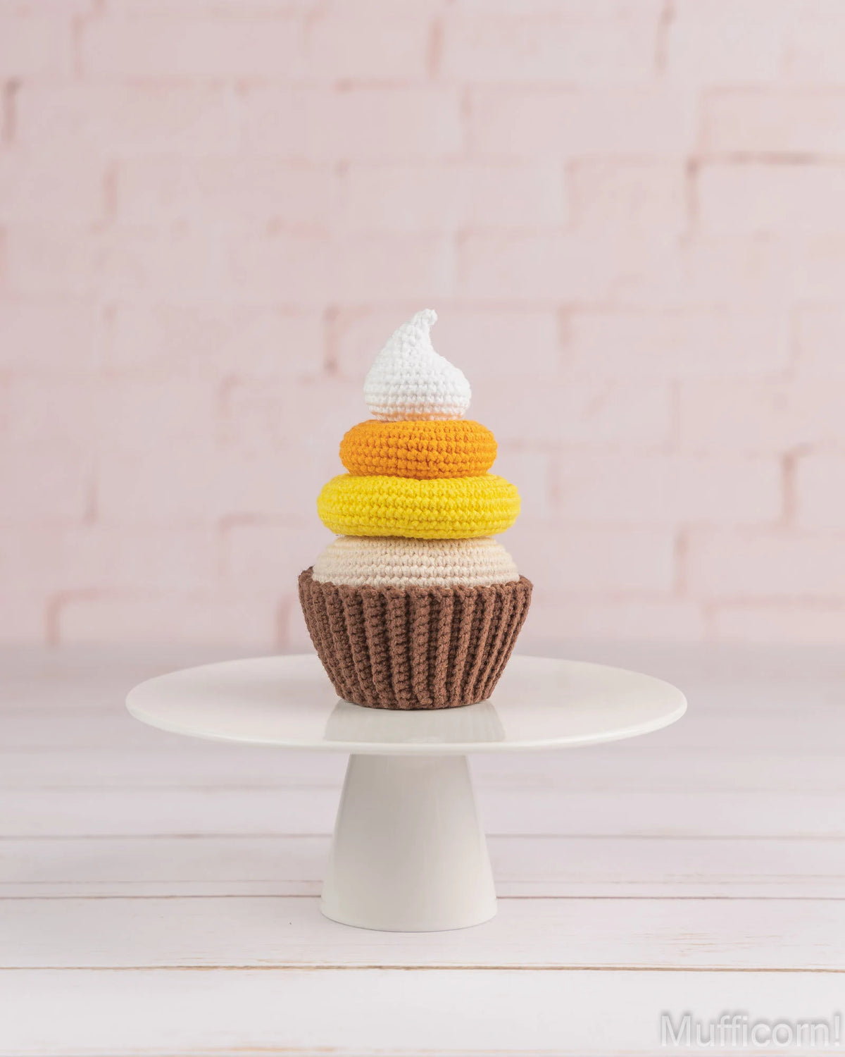 A crocheted cupcake decoration or plush toy with a brown base, yellow, orange, and cream layers, and a white frosting top, displayed on a white pedestal stand against a pale pink brick wall.