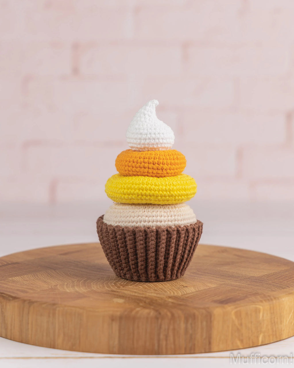 A crocheted cupcake decoration or small cupcake figure sits on a round wooden board against a soft pink brick wall background. The figure has a white top, then white, yellow, and orange frosting-like layers, with a brown base.
