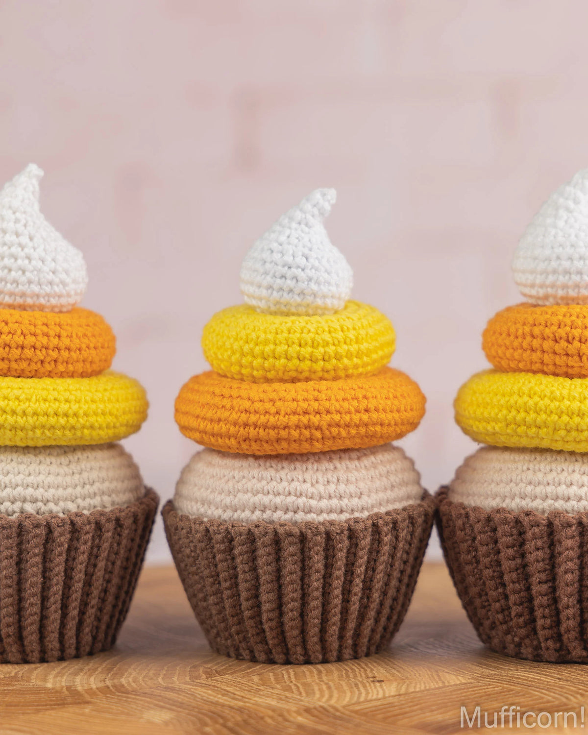 Three crocheted cupcake-shaped ornaments displayed on a wooden surface against a soft pink background. Each cupcake has a brown textured base, yellow and orange rings, a cream-colored middle band, and a white frosting-like top.