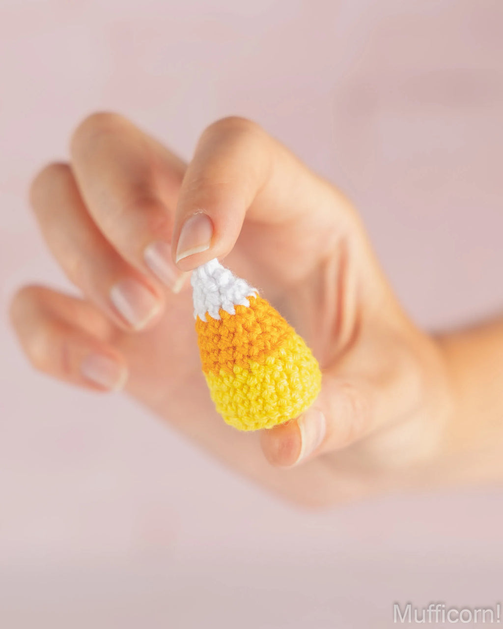 A hand holding a small crocheted candy corn accessory with yellow, orange, and white sections against a soft pink background.