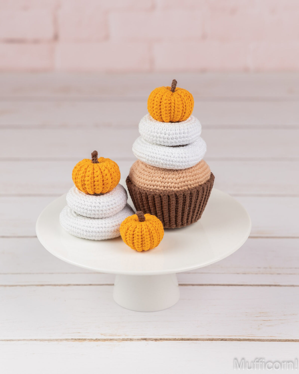 Three crocheted cupcake ornaments with orange pumpkins on top, displayed on a white cake stand against a light pink brick wall and white wood background.