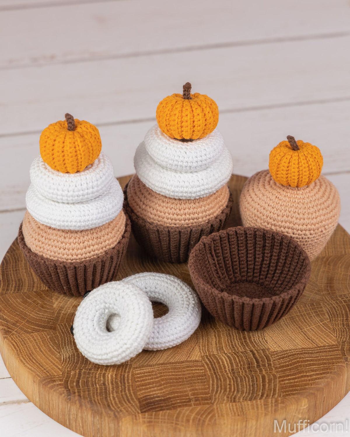 Three crocheted cupcake ornaments with orange pumpkins on top, displayed on a round wooden tray with two white donut ornaments and a brown cupcake liner against a white wood plank background.