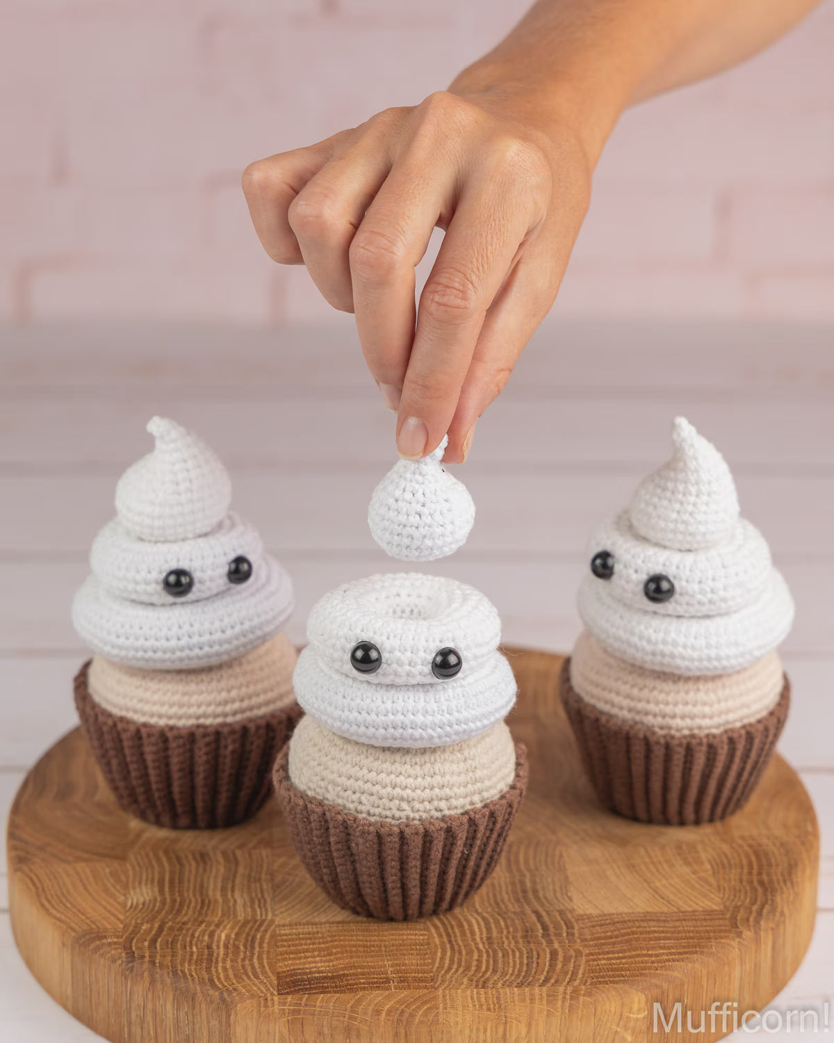 A hand places a small white crocheted ghost-shaped decoration on top of a white and beige crocheted poop cupcake, with two matching cupcakes on a round wooden board against a light pink brick wall.