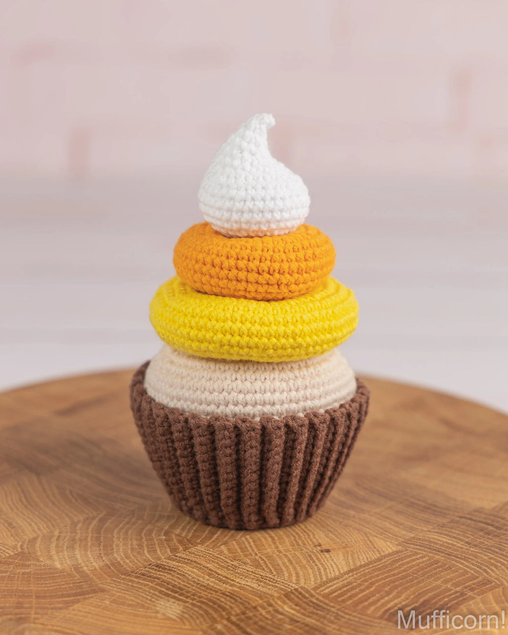 A crocheted cupcake toy with a brown base, cream, yellow, orange, and white frosting layers, displayed on a round wooden surface against a soft pink background.