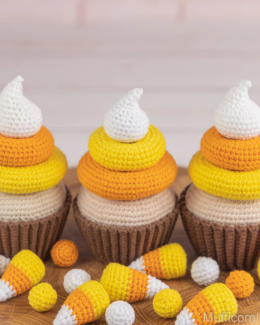 Three crocheted cupcakes styled like candy corn with brown paper liners, cream, yellow, orange, and white crochet layers, with small loose candy corn pieces scattered in front on a wooden surface.
