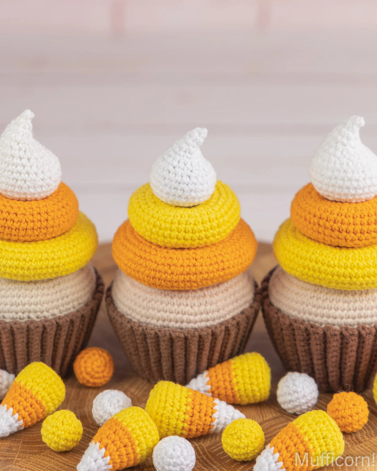 Three crocheted cupcakes styled like candy corn with brown paper liners, cream, yellow, orange, and white crochet layers, with small loose candy corn pieces scattered in front on a wooden surface.