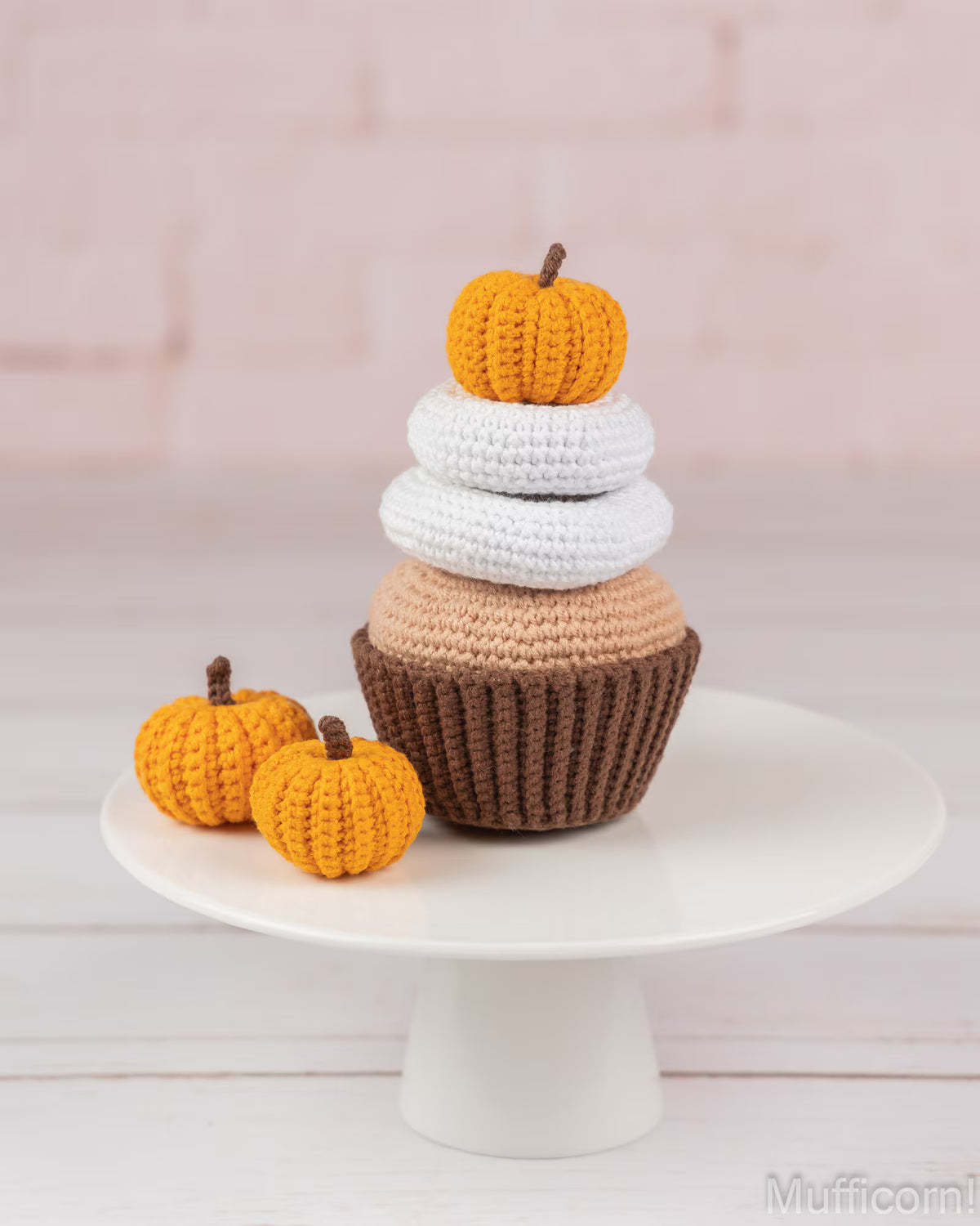 A crocheted pumpkin cupcake decoration displayed on a white cake stand, featuring a brown base, white frosting, a beige filling layer, and two small orange pumpkins on top, set against a soft pink brick wall background.