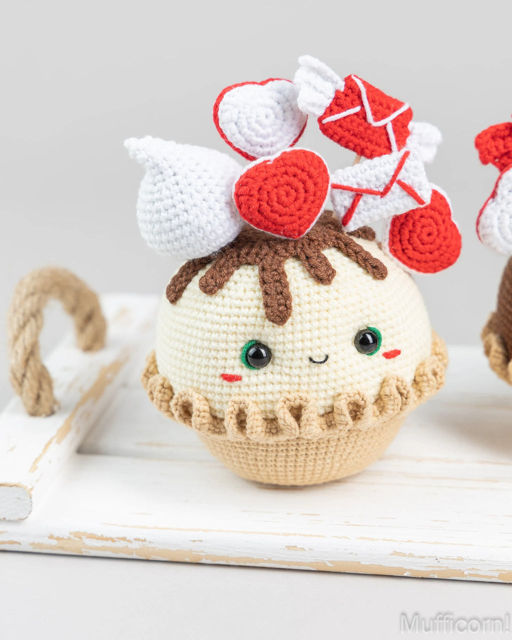 A cute crocheted plush toy shaped like a cupcake or dessert, with cream-colored frosting, brown yarn hair, and red-and-white heart-shaped accessories on top. It is photographed on a white rustic surface against a light gray background.