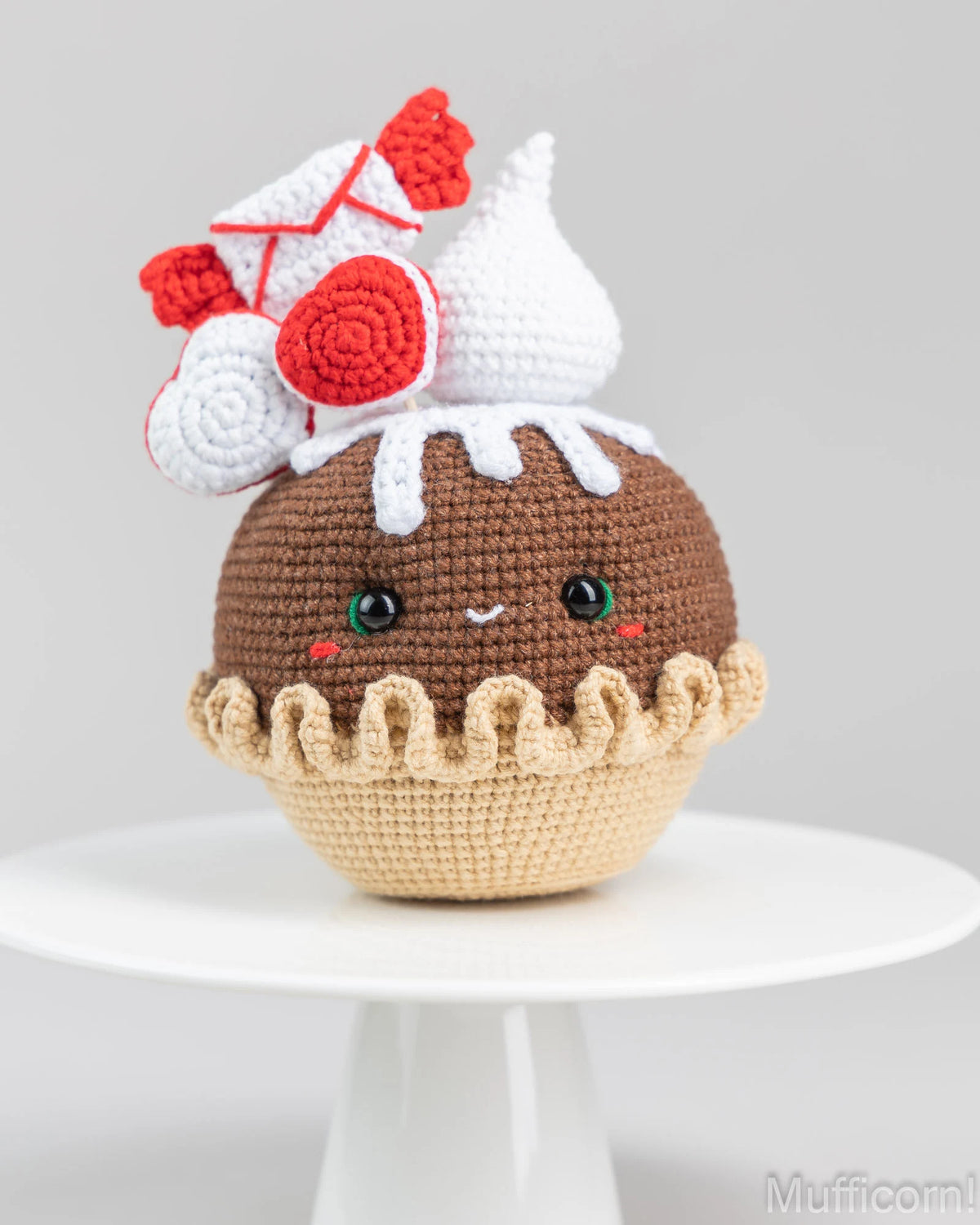 A crocheted plush toy shaped like a cupcake or dessert, sitting on a white pedestal against a light gray background. The toy has a brown body, a beige ruffled base, white frosting details, and red accents on the top.