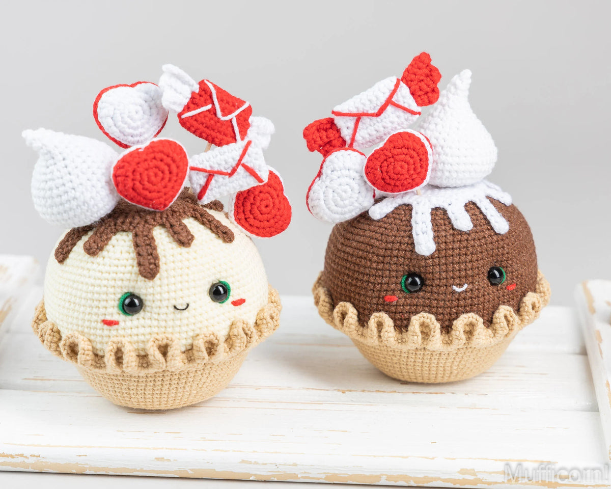 Two cute crocheted dessert dolls, one resembling a vanilla pie with red heart-shaped decorations and one resembling a chocolate cake with white frosting and red heart decorations, displayed on a white wooden surface against a light gray background.