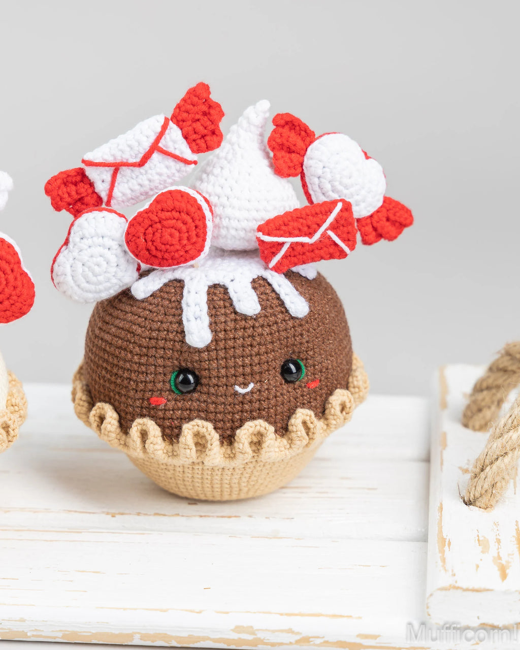 A cute crocheted plush toy shaped like a chocolate cupcake or dessert, with a brown body, beige crimped base, white frosting-like top, and red heart and envelope decorations. It is photographed on a white wooden surface against a soft light gray background.