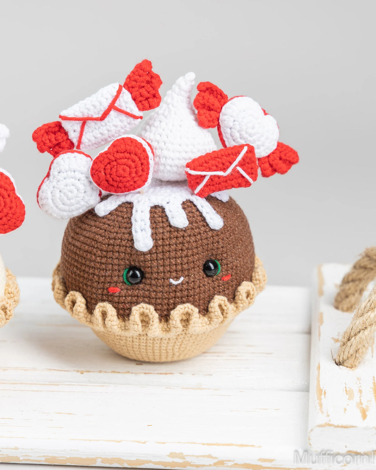 A cute crocheted plush toy shaped like a chocolate cupcake or dessert, with a brown body, beige crimped base, white frosting-like top, and red heart and envelope decorations. It is photographed on a white wooden surface against a soft light gray background.