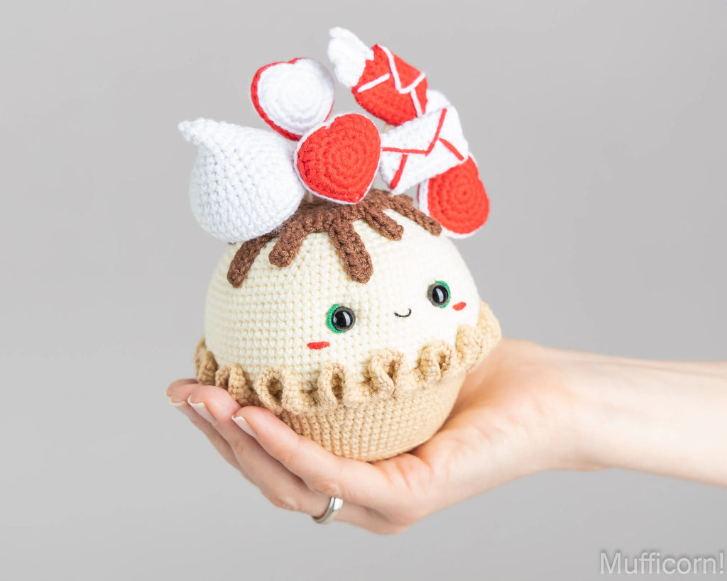 A hand holds a cute crocheted cupcake plush toy with a smiling face, cream-colored body, brown frosting, and red and white whipped cream toppings against a plain gray background.