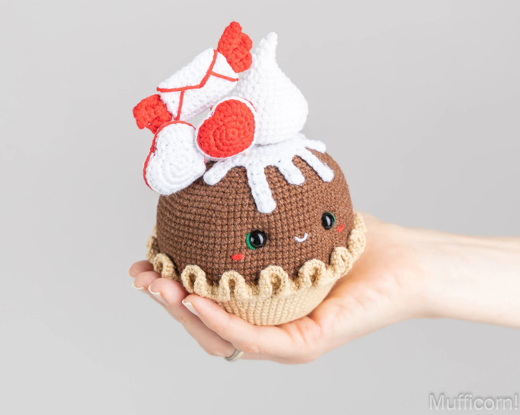 A hand holding a crocheted cupcake toy with a brown body, beige base, white frosting, and red heart-shaped decorations on a light gray background.