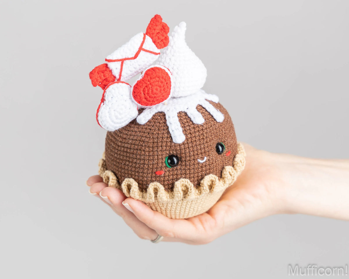 A hand holding a crocheted cupcake toy with a brown body, beige base, white frosting, and red heart-shaped decorations on a light gray background.