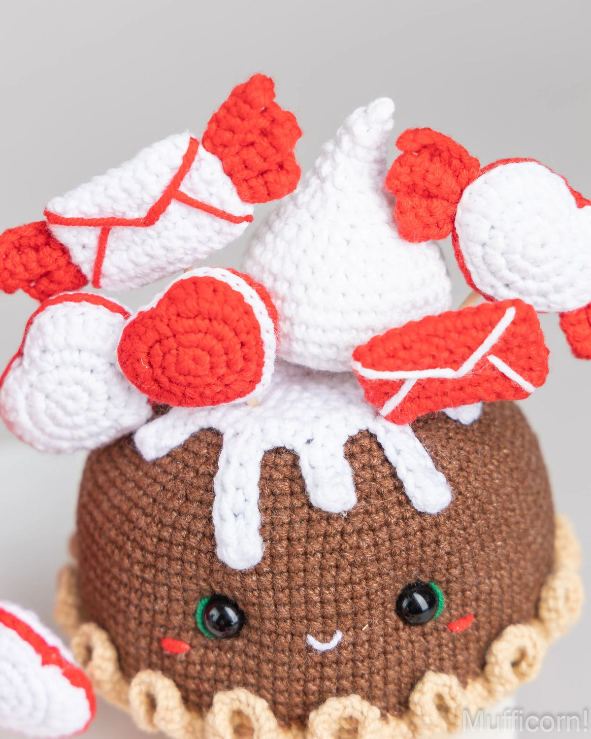 A close-up of a crocheted amigurumi character resembling a chocolate cake or dessert, with white frosting-like detailing, a white cone on top, and red-and-white candy motifs. The face has black eyes and a small smile.