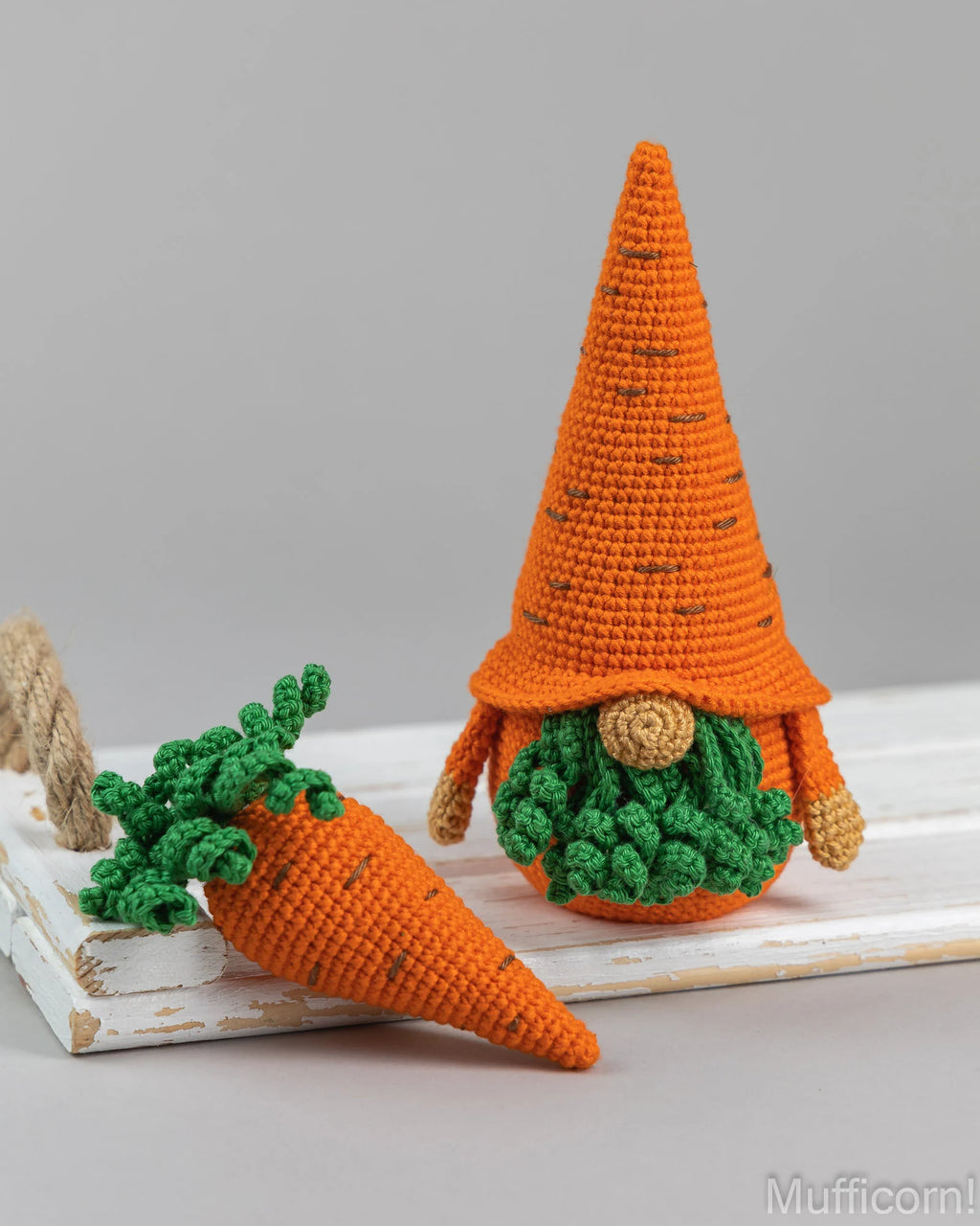 Crocheted carrot gnome doll with an orange pointed hat, green leafy top, and round green body, displayed beside a matching crocheted carrot on a white wooden surface against a light gray background.