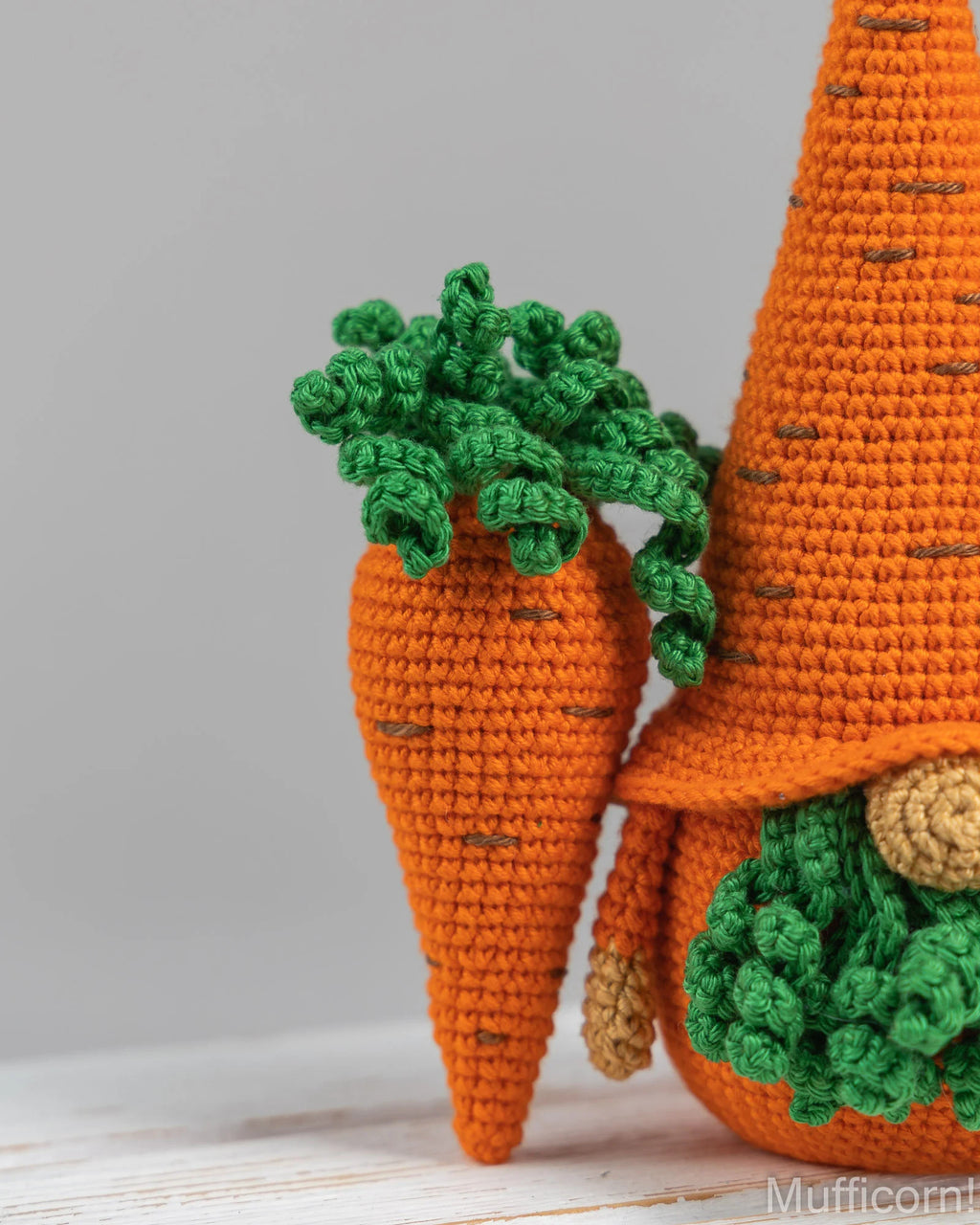 A close-up of a crocheted carrot gnome decoration, showing an orange textured body, green leafy top, and part of a larger matching carrot gnome behind it on a light gray background.