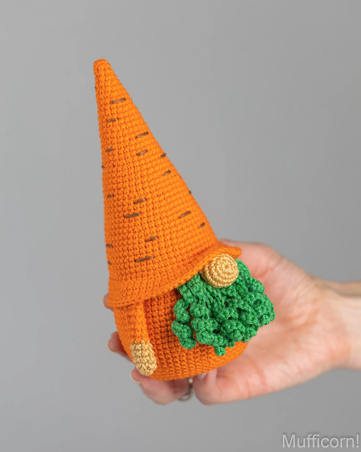 A hand holds a crocheted carrot gnome with a pointed orange hat, green leafy details, and small tan accents against a plain gray background.