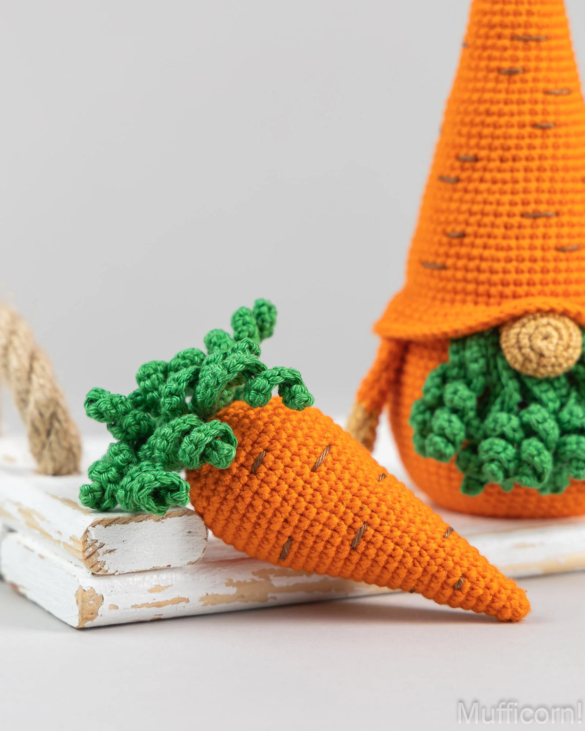 Crocheted carrot-shaped gnome decoration in orange and green, shown leaning on stacked white wooden blocks with a second carrot gnome visible in the background against a light gray studio backdrop.