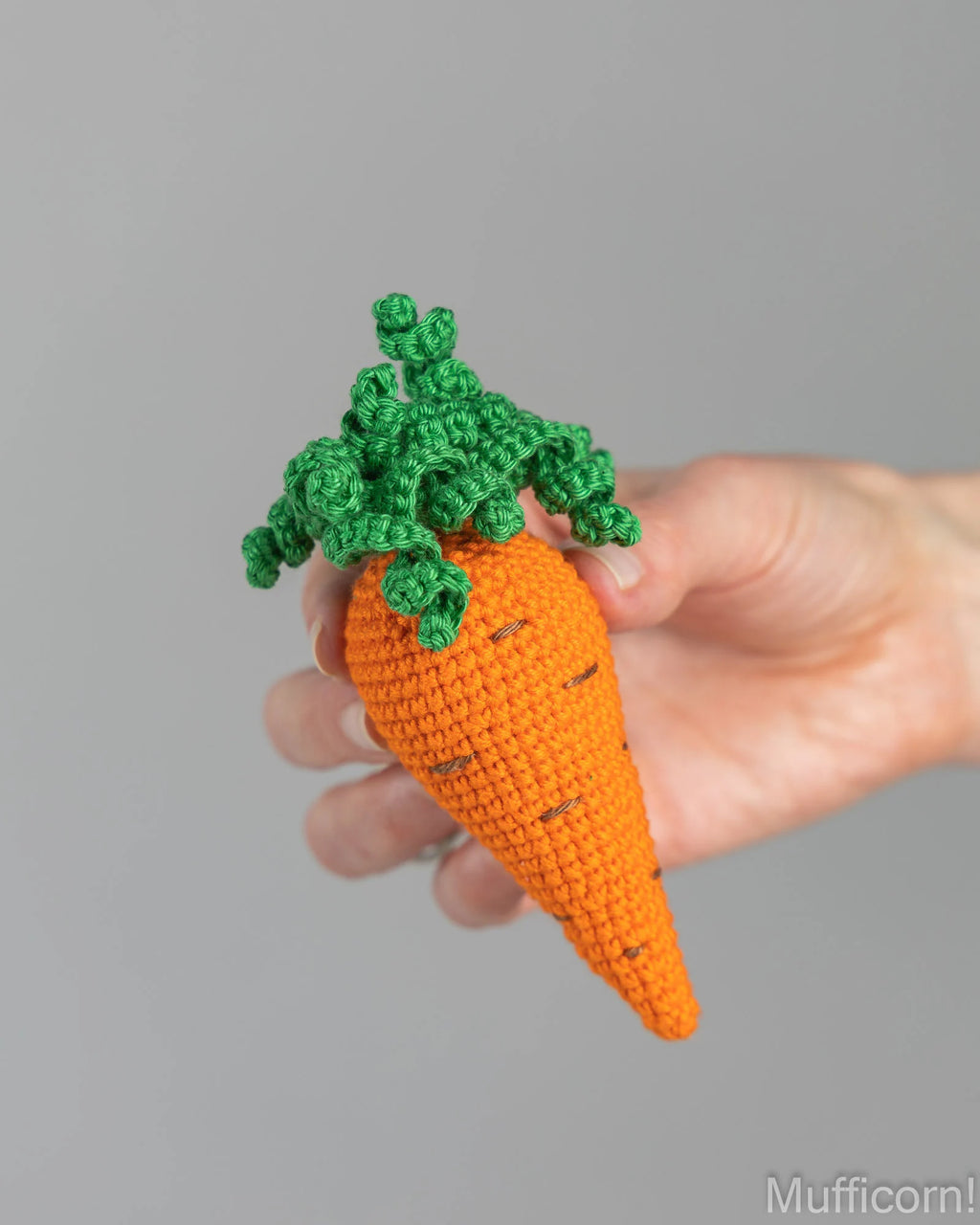 A crocheted carrot ornament with a bright orange body and green leafy top, held in a person's hand against a plain gray background.