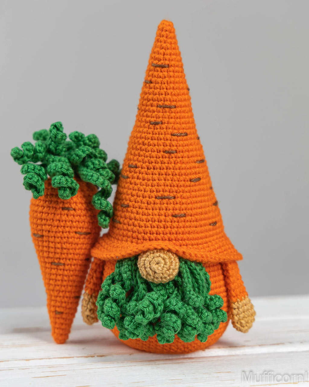 A crocheted carrot gnome doll with a tall orange pointed hat, green leafy top, and round tan face, standing beside another smaller carrot gnome on a light surface against a soft gray background.
