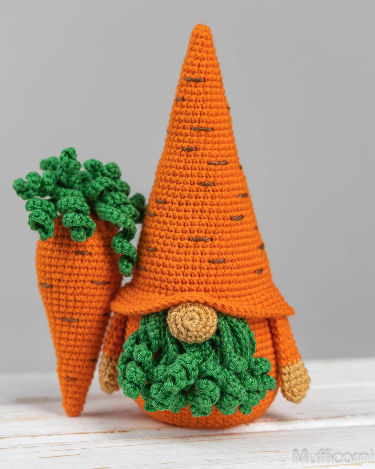 A crocheted carrot gnome doll with a tall orange pointed hat, green leafy top, and round tan face, standing beside another smaller carrot gnome on a light surface against a soft gray background.