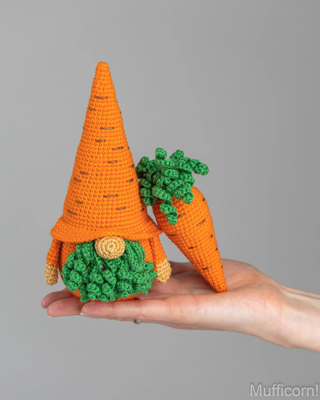 A crocheted carrot gnome with a tall orange hat, green leafy top, and round green beard is being held in a person's hand against a plain gray background.