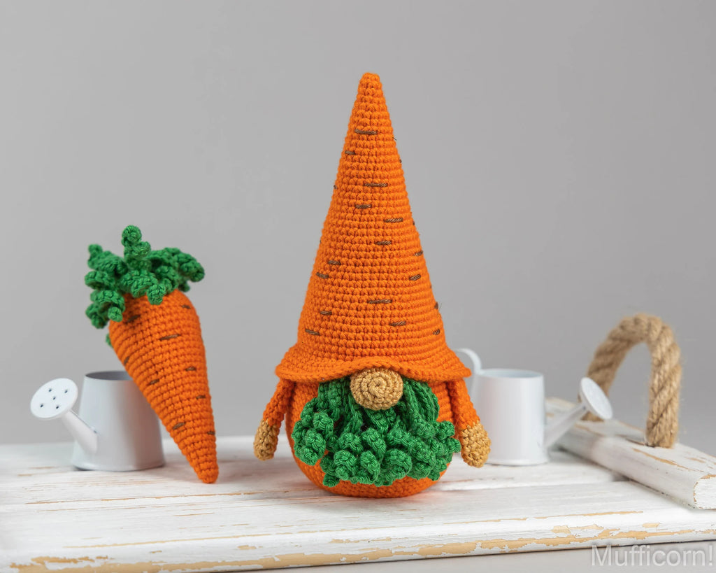 Crocheted carrot-shaped gnome decoration and a matching carrot topper, displayed on a white wooden surface with small white watering cans against a light gray background.